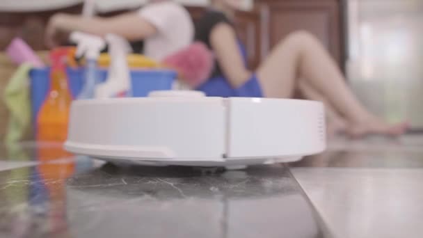 Young tired man and woman sitting on the floor back to back on the background. Cleaning day. Cleaning equipment is nearby. The robot vacuum cleaner moving on the floor close-up. Bottom view — Stockvideo