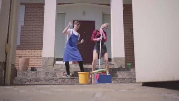 Cute young woman and man dancing on the porch of the house. Couple cleaning house together. Happy routine life. Housekeeping household housework and cleaning service concept. Slow motion. — Stock Video