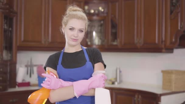 Retrato linda mujer joven de pie en la cocina con las manos cruzadas sosteniendo botellas de detergente, mirando a la cámara sonriendo. Una ama de llaves positiva limpiando su casa. El concepto de — Vídeo de stock