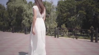 Uzun esmer saçlı büyüleyici genç kız güneş gözlüğü ve uzun beyaz yaz moda elbise sokakta yürürken giyiyor. Yumuşak gün batımı doğal ışık ile parkta güzel bir kadının eğlence.