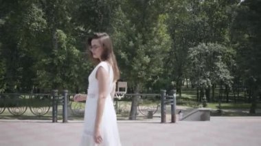 Uzun esmer saçlı sevimli genç kız güneş gözlüğü ve uzun beyaz yaz moda elbise sokakta yürürken giyiyor. Yumuşak gün batımı doğal ışık ile parkta güzel bir kadının eğlence.