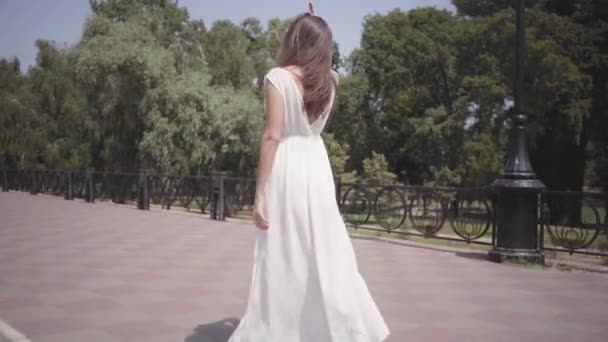 Όμορφο νεαρό κορίτσι φορώντας γυαλιά ηλίου και ένα μακρύ λευκό καλοκαιρινό φόρεμα μόδας με τα πόδια μέσα από την πόλη. Αναψυχή από μια όμορφη γυναίκα στο πάρκο με απαλό ηλιοβασίλεμα φυσικό φως. — Αρχείο Βίντεο