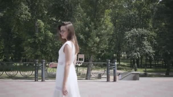 Cute młoda dziewczyna z długim Brunetka włosy noszenie okulary przeciwsłoneczne i długie białe lato mody sukienka chodzenie w dół ulicy. Wypoczynek pięknej kobiety w parku z miękkim zachodzie słońca naturalnego światła. — Wideo stockowe