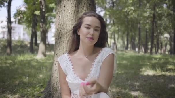 Portrait jeune fille glamour avec de longs cheveux bruns portant une longue robe de mode d'été blanche assise sous un arbre dans le parc. Loisirs d'une jolie femme regardant la caméra à l'extérieur . — Video