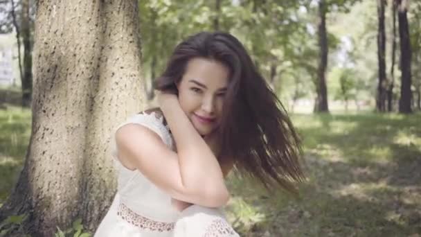 Portrait adorable jeune fille aux longs cheveux bruns portant une longue robe de mode blanche d'été assise sous un arbre dans le parc. Loisirs d'une jolie femme regardant la caméra à l'extérieur . — Video