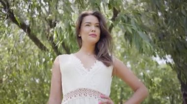 Uzun esmer saçlı portre güzel genç kız parkta bir ağacın dalları altında duran uzun beyaz yaz moda elbise giyiyor. Boş zaman güzel kadın kamera açık havada bakıyor.