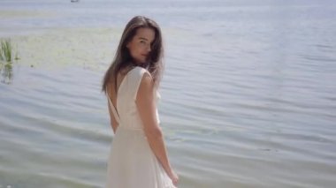 Uzun esmer saçlı portre büyüleyici genç kız nehir yanında duran uzun beyaz yaz moda elbise giyiyor. Boş zaman güzel kadın kamera açık havada bakıyor.