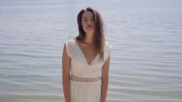Retrato hermosa joven con el pelo largo morena usando un vestido largo de moda de verano blanco de pie junto al río. Ocio mujer bonita mirando a la cámara al aire libre . — Vídeos de Stock