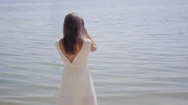 Portret glamoureuze jong meisje met lange brunette haar dragen een lange witte zomer mode jurk staande aan de rivier. Vrije tijd mooie vrouw kijken naar de camera buitenshuis. — Stockvideo