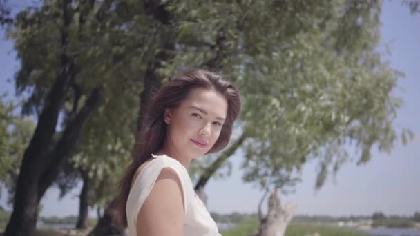 Portret mooi jong meisje met lange brunette haar dragen een lange witte zomer mode jurk staande aan de rivier. Vrije tijd mooie vrouw kijken naar de camera buitenshuis. — Stockvideo