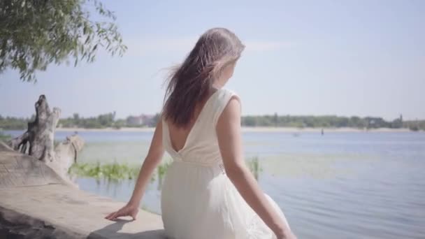 Portret efektowna młoda dziewczyna z długimi brunetką włosy ubrana w długą białą sukienkę mody siedzącej na nabrzeżu. Rozrywka ładna kobieta patrząc na kamerę na zewnątrz. — Wideo stockowe