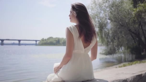 Portrait charmante jeune fille avec de longs cheveux bruns portant une longue robe blanche de mode d'été assis sur le front de mer. Loisirs jolie femme en plein air . — Video