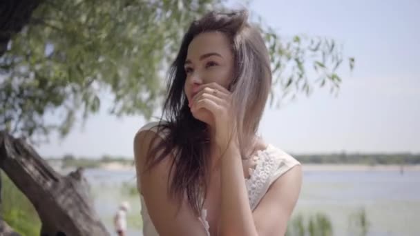 肖像迷人的年轻女孩与长黑发穿着长长的白色夏季时尚礼服坐在海滨。休闲漂亮的女人看着相机拉直头发户外. — 图库视频影像