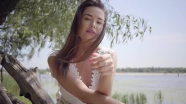 Uzun esmer saçlı portre sevimli genç kız rıhtımda oturan uzun beyaz yaz moda elbise giyiyor. Boş zaman güzel kadın açık havada saç doğrultma kamera bakıyor.