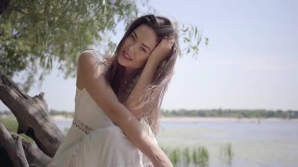 Portrait charmante jeune fille avec de longs cheveux bruns portant une longue robe blanche de mode d'été assis sur le front de mer. Loisirs jolie femme regardant la caméra redresser les cheveux à l'extérieur . — Video