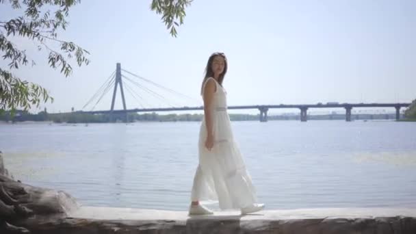 Muchacha joven glamorosa retrato con el pelo largo morena usando un vestido largo de moda de verano blanco caminando en el paseo marítimo. Ocio mujer bonita mirando a la cámara al aire libre . — Vídeo de stock