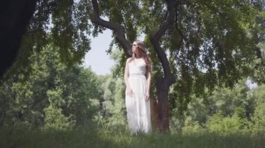 Uzun esmer saçlı portre sevimli genç kız güneş gözlüğü ve parkta bir ağacın dalları altında duran uzun beyaz yaz moda elbise giyiyor. Boş güzel bir kadın açık havada.
