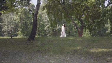 Parkta bir ağacın dalları altında duran uzun beyaz yaz moda elbise giyen uzun esmer saçlı sevimli genç kız. Boş güzel bir kadın kamera açık havada bakıyor.