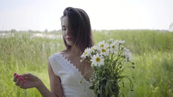 肖像可爱的年轻女孩与黑发穿着长长的白色夏季时尚礼服站在田野上。休闲漂亮的女人拿着一束洋甘菊看着户外的相机. — 图库视频影像