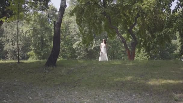 Aranyos fiatal lány, hosszú barna hajú visel, hosszú, fehér nyári divat ruha alatt álló ágak egy fa a parkban. Szabadidő egy csinos nő nézi a kamerát a szabadban. — Stock videók