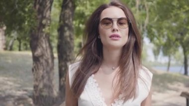 Güneş gözlüğü ve parkta ahşap bir köprü boyunca yürüyen uzun beyaz yaz moda elbise giyen esmer saçlı Portre güzel genç kız. Boş güzel bir kadın kamera açık havada bakıyor.