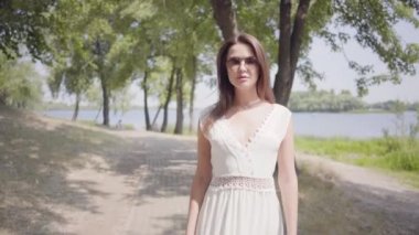 Güneş gözlüğü ve park boyunca yürüyen uzun beyaz yaz moda elbise giyen esmer saçlı Portre çekici genç kız. Boş güzel bir kadın kamera açık havada bakıyor.