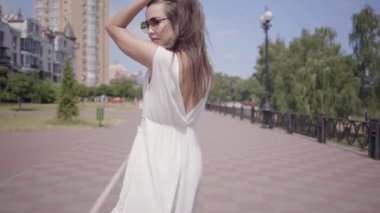 Güneş gözlüğü ve açık havada yürüyen uzun beyaz yaz moda elbise giyen sevimli genç kız. Güzel bir kadının boş boş boş kameraya bakmak etrafında döner. Yavaş çekim.