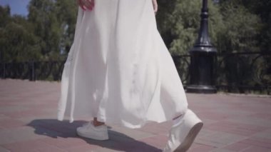Tanınmayan kız açık havada yürüyen uzun beyaz yaz moda elbise giyiyor. Güzel bir kadının boş zamanları. Yavaş çekim.
