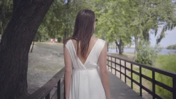 Retrato de chica joven con cabello moreno con gafas de sol y vestido largo de verano blanco caminando a lo largo del parque. Ocio una mujer bonita al aire libre . — Vídeos de Stock