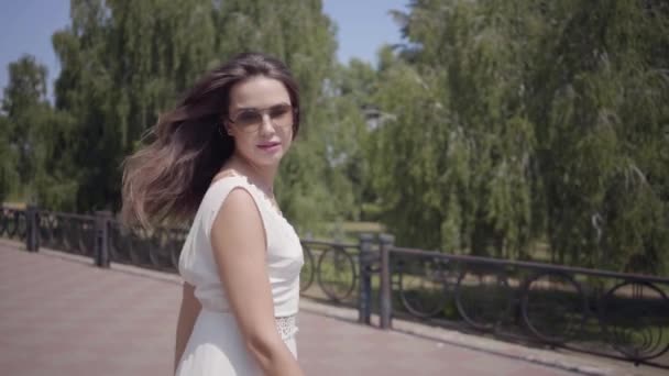 Encantadora joven con gafas de sol y un largo vestido de verano blanco de moda caminando al aire libre. Ocio de una mujer bonita se da la vuelta alisando el cabello y mirando a la cámara. Movimiento lento . — Vídeo de stock