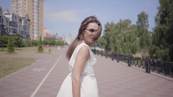 Chica joven glamurosa con gafas de sol y un vestido largo y blanco de verano caminando al aire libre. Ocio de una mujer bonita se da la vuelta mirando a la cámara. Movimiento lento . — Vídeo de stock