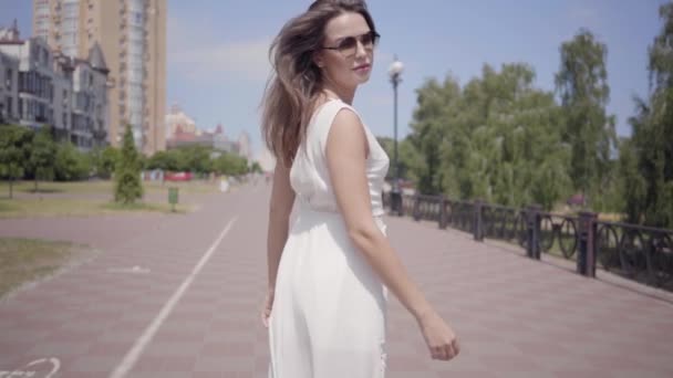 Charming young girl wearing sunglasses and a long white summer fashion dress walking outdoors. Leisure of a pretty woman turns around lookng into the camera. Slow motion. — Stock Video