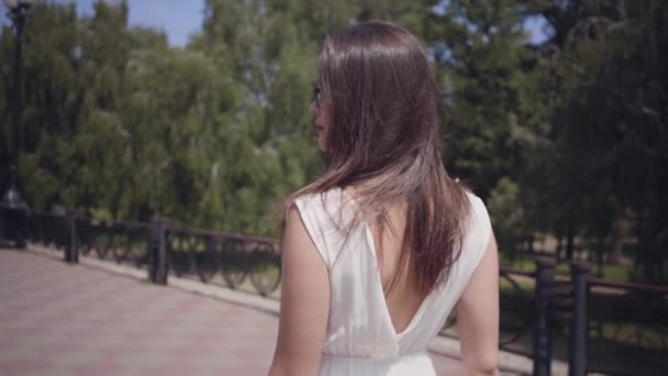 Muchacha joven glamorosa retrato con gafas de sol y un vestido largo de moda de verano blanco caminando al aire libre. Ocio de una mujer bonita en el parque. Movimiento lento . — Vídeo de stock