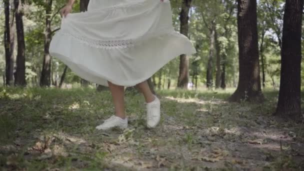 Chica joven no reconocida vestida con un vestido blanco de verano largo gira en el parque. Ocio de una mujer bonita al aire libre. Movimiento lento . — Vídeos de Stock