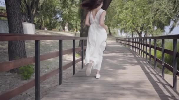 Muchacha joven adorable retrato con pelo moreno con gafas de sol y vestido de verano largo y blanco corriendo sobre un puente de madera. Ocio a una mujer bonita al aire libre. Movimiento lento . — Vídeos de Stock