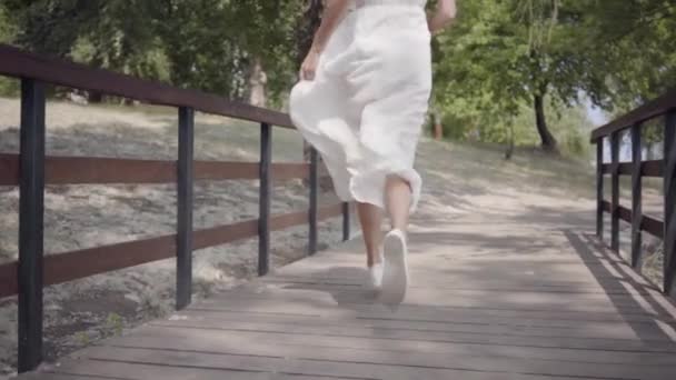 Portret schattig jong meisje met brunette haar dragen zonnebril en lange witte zomer mode jurk loopt over een houten brug. Vrije tijd een mooie vrouw buitenshuis. Slow Motion. — Stockvideo