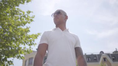 Güneş gözlüğü ve beyaz t-shirt giyen emin başarılı kel orta doğu adam. Yakışıklı iş adamı bir şehir caddesinde güneşin üzerinde duran etrafında bakar. Eğlence moda adam.