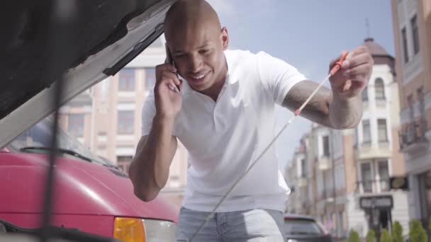 Back view of bald middle eastern man looking under the car hood speaking by the cell phone. The guy checking the broken car outdoors. Automobile fixing. — Stock Video