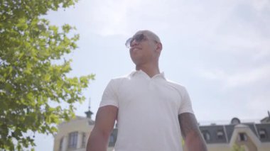 Portre emin başarılı gülümseyen kel orta doğu adam güneş gözlüğü ve beyaz t-shirt giyiyor. Yakışıklı iş adamı bir şehir caddesinde güneşin üzerinde duran etrafında bakar. Eğlence moda adam.