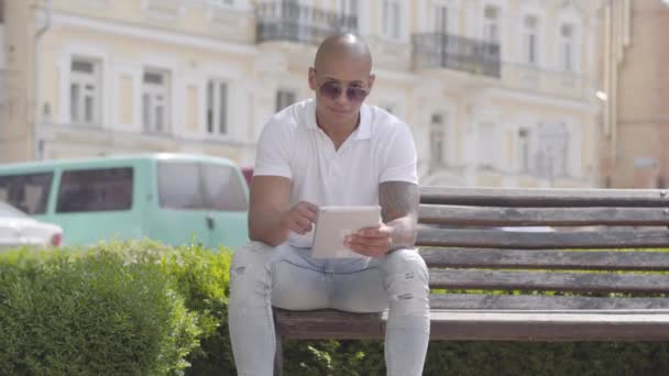 Hombre guapo calvo de Oriente Medio con gafas de sol y camiseta blanca escribiendo en la tableta sentada en el banco en la calle. El tipo que pasa tiempo al aire libre. Concepto independiente . — Vídeos de Stock