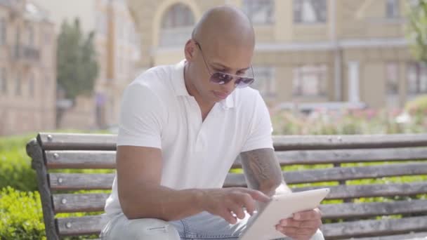 Portrait handsome bald middle eastern man wearing sunglasses and white t-shirt with tablet sitting on the bench on the street. The guy spending time outdoors. Freelance concept. — Stock Video