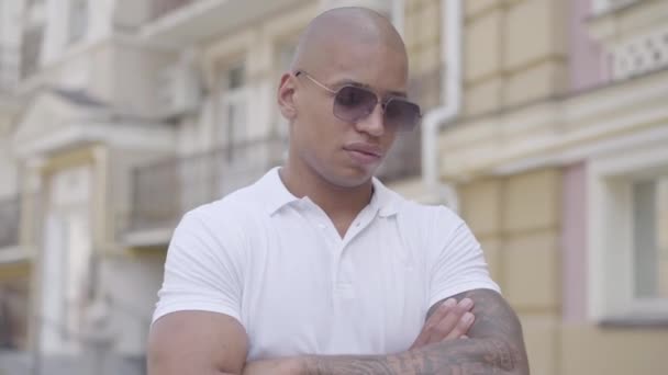 Portrait of handsome confident bald middle eastern man wearing sunglasses and white t-shirt with crossed arms looking in the camera standing on the street in front of old buildings. — Stock Video