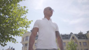Güneş gözlüğü ve beyaz t-shirt giyen emin başarılı kel orta doğu adam. Yakışıklı iş adamı bir şehir caddesinde güneşin üzerinde duran etrafında bakar. Boş zaman moda adam. Yavaş çekim.