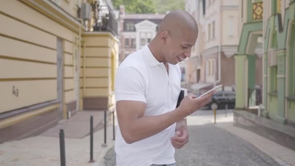 Retrato de belo e bem-sucedido careca confiante médio oriental digitando no telefone celular em pé na rua em frente a edifícios antigos olhando ao redor. Competências profissionais . — Vídeo de Stock