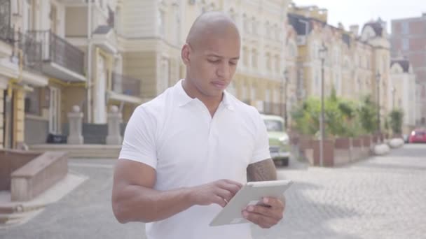 Retrato de belo homem calvo e confiante do Oriente Médio digitando no tablet em pé na rua em frente a velhos e bonitos edifícios. Competências profissionais, processo de trabalho . — Vídeo de Stock