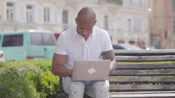 Portre yakışıklı kel orta doğu işadamı sokakta bankta oturan dizüstü bilgisayar ile çalışıyor. Serbest kavram. Dışarıda çalışan bir adam. Yavaş çekim. — Stok video