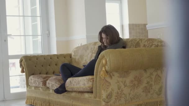 Молодая красивая девушка сидит и расслабляется со столиком на диване в уютной гостиной и весело улыбается камере — стоковое видео