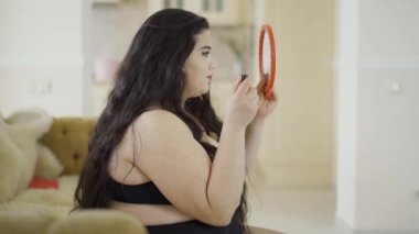 Aynada bakarak makyaj yapıyor çekici genç artı boyutu kız Portresi. Parlak kırmızı ruj ile dudaklarını boyama tombul kadın yan görünümü. Tarihe hazırlanıyorum. Üfleme öpücük bakarak