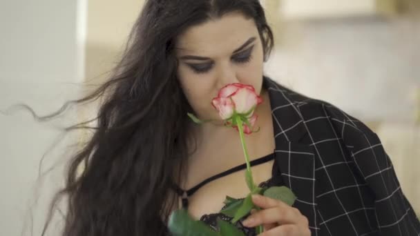 Portrait of adorable overweight woman sniffing rose indoors. The hair fluttering on the wind. A sensual plump young woman looking at camera. Tenderness, sensuality. — Stock Video