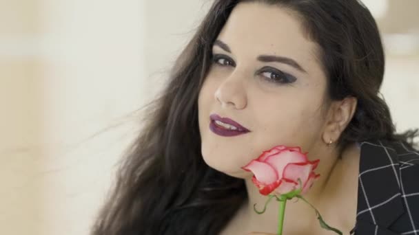 Ritratto ravvicinato di attraente ragazza plus size in una giacca nera annusare rosa. Una giovane donna grassoccia sensuale con un trucco luminoso guardando nella fotocamera . — Video Stock